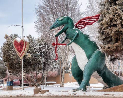 A large green dragon statue with wings stands in a snowy park, next to a heart-shaped decoration.