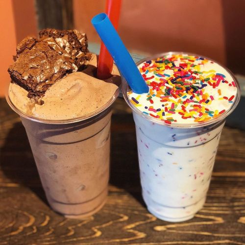 Two milkshakes in clear cups: one chocolate topped with a brownie, the other vanilla with colorful sprinkles.