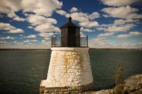 A stone lighthouse stands on a rocky shore, surrounded by water under a blue sky with fluffy clouds.