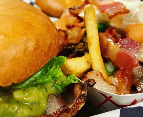 A close-up of a burger with lettuce and guacamole, alongside a pile of fries topped with bacon bits.
