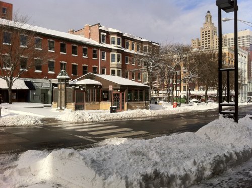 A snowy street scene featuring brick buildings, a bus stop, and snow piled along the sidewalks.