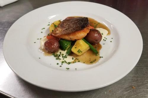 A plated dish featuring seared fish atop colorful vegetables and a light sauce, garnished with herbs.
