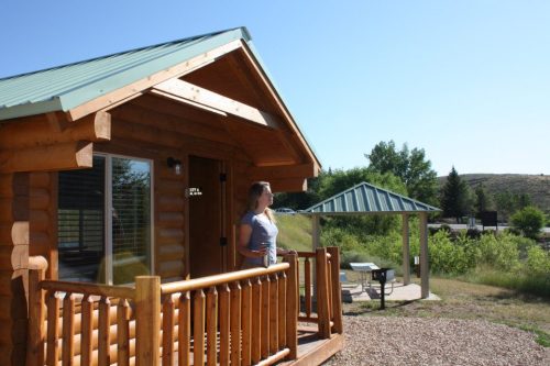 A woman stands on the porch of a log cabin, surrounded by greenery and a picnic area under a blue sky.