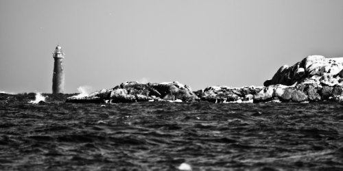 A black and white image of a lighthouse in the distance, surrounded by rocky shores and turbulent ocean waves.
