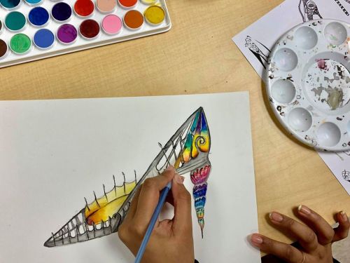 A person paints a colorful high-heeled shoe design with watercolors on a white sheet, surrounded by paint palettes.