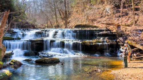 A serene waterfall cascades over rocks, with a person standing by the water's edge in a wooded area.
