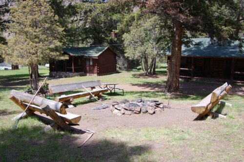 A campsite with a fire pit surrounded by log benches and cabins in a wooded area.
