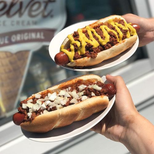 Two hands hold plates with large hot dogs: one topped with mustard and chili, the other with chili and onions.