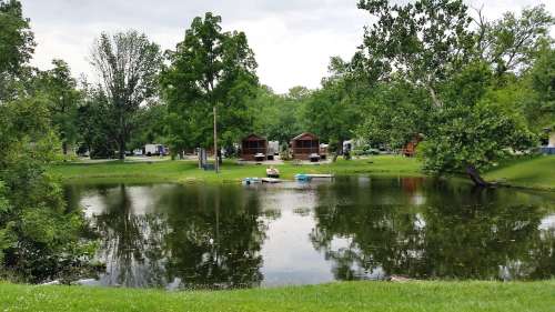 A serene pond surrounded by trees, with two cabins in the background and a small boat on the water.