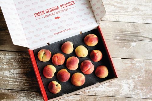 A box of fresh Georgia peaches, neatly arranged on black foam, with a rustic wooden background.