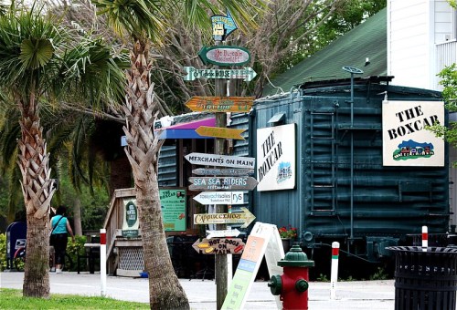 A collection of colorful directional signs in front of a green train car, surrounded by palm trees and a sidewalk.