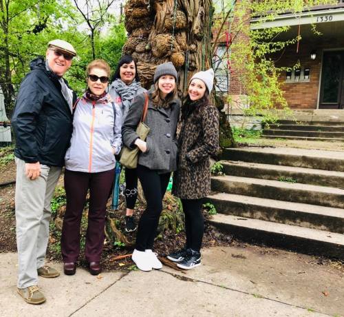A group of five people poses together outdoors near a tree and steps, smiling and dressed for cool weather.