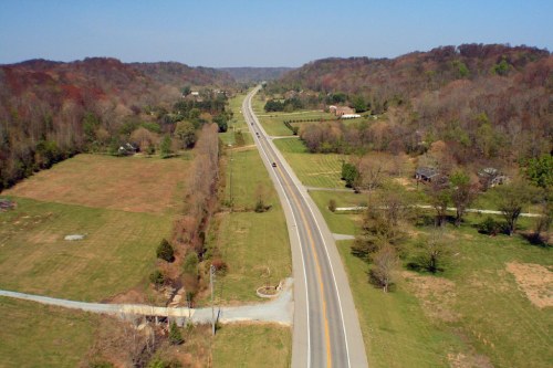Aerial view of a winding road through green fields and trees, with distant hills under a clear blue sky.