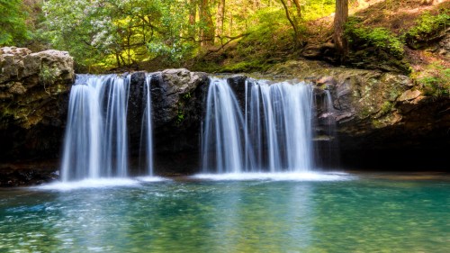 A serene waterfall cascades over rocks into a clear, tranquil pool surrounded by lush greenery.