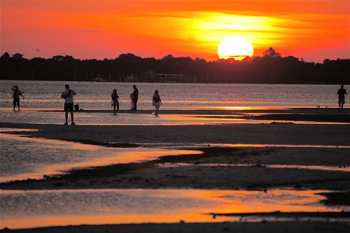 Silhouettes of people walking along a beach at sunset, with vibrant orange and yellow hues reflecting on the water.