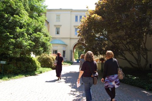 Three people walking down a brick path towards a large building, surrounded by greenery and trees.