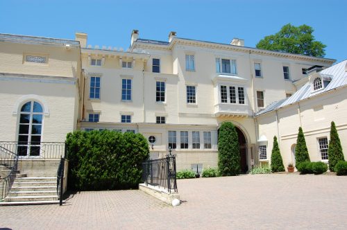 A large, historic building with a light-colored facade, surrounded by greenery and a paved courtyard under a clear blue sky.