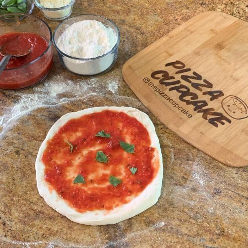 A pizza dough topped with tomato sauce and fresh basil, with flour and sauce in bowls nearby, on a wooden cutting board.