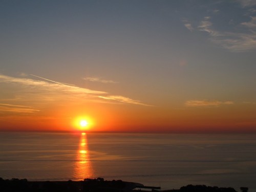 A vibrant sunset over calm waters, with orange and yellow hues reflecting on the surface and wispy clouds in the sky.