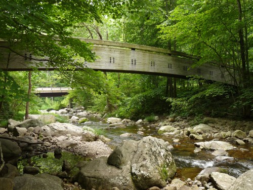 A wooden bridge arches over a serene stream, surrounded by lush green trees and rocky riverbanks.
