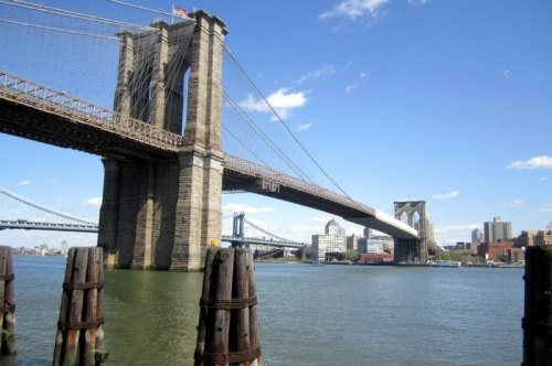 Brooklyn Bridge spanning a river, with city skyline and another bridge visible in the background under a clear blue sky.