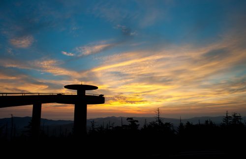 Silhouette of a viewing platform against a colorful sunset sky with mountains in the background.
