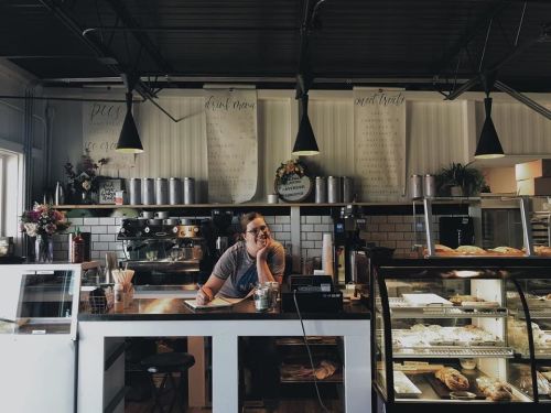 A barista smiles at the counter of a cozy café, surrounded by pastries and coffee equipment.