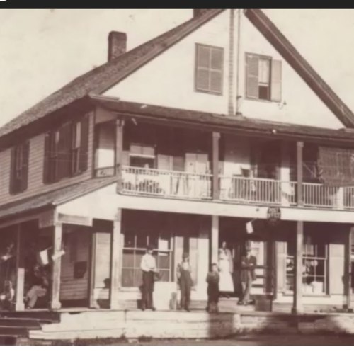 Historic black-and-white photo of a two-story house with a porch, featuring several people standing outside.