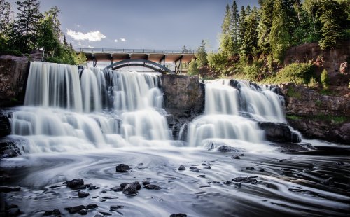 A serene waterfall cascades over rocks, with a bridge in the background surrounded by lush greenery and a clear sky.