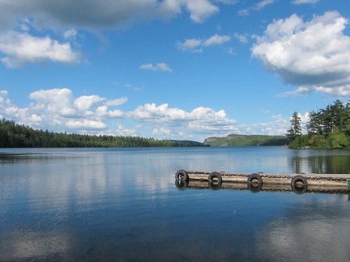 A serene lake scene with a wooden dock, surrounded by lush greenery and fluffy clouds in a blue sky.