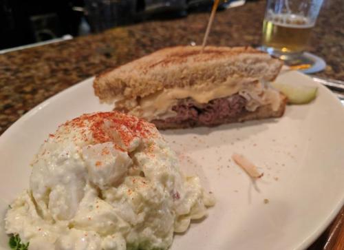A plate with a sandwich filled with meat and sauerkraut, alongside a serving of creamy potato salad sprinkled with paprika.