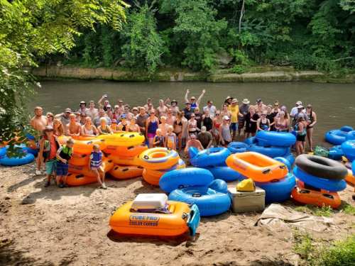 A large group of people gathered by a river, surrounded by colorful inflatable tubes on the sandy shore.