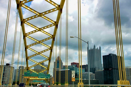 View from a bridge with yellow cables, showcasing Pittsburgh's skyline and cloudy skies in the background.