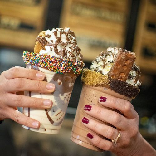 Two hands hold colorful milkshakes topped with whipped cream, sprinkles, and chocolate bars against a blurred background.