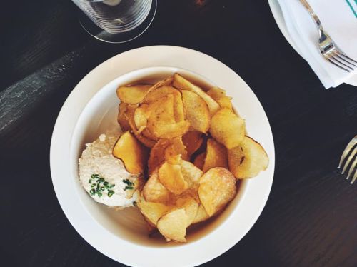 A bowl of crispy potato chips served with a creamy dip, garnished with chives, on a dark wooden table.