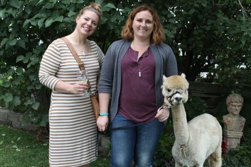 Two women stand beside an alpaca in a garden, one holding a drink and the other holding the alpaca's leash.