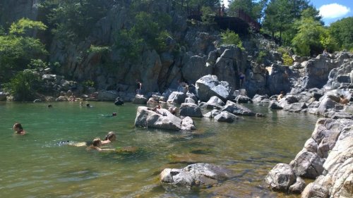 People swimming and relaxing in a natural pool surrounded by rocky cliffs and greenery on a sunny day.