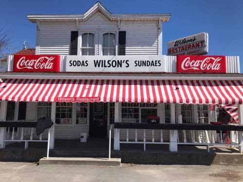 A vintage restaurant with a striped awning, featuring signs for Coca-Cola and "Wilson's Sundaes" on the facade.