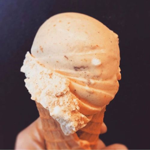 A hand holding a cone with two scoops of creamy ice cream, one light brown and one white, against a dark background.
