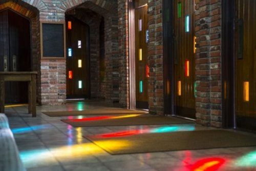 A hallway with brick walls and colorful light reflections on the floor from stained glass windows.