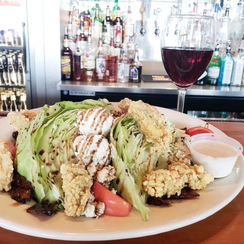 A plate of salad with crispy toppings, tomatoes, and a side of dressing, accompanied by a glass of red wine.