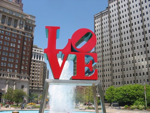 A large red sculpture of the word "LOVE" stands in a city plaza with buildings and a fountain in the background.