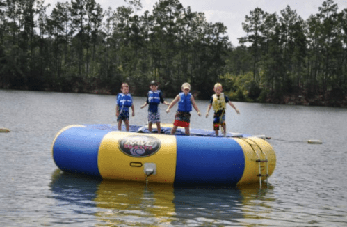 Four children in life jackets stand on a large inflatable water trampoline in a lake, surrounded by trees.