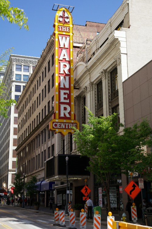 The Warner Center marquee stands tall on a sunny street, surrounded by trees and construction signs.