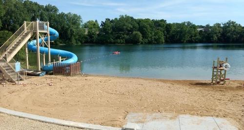 A sandy beach with a blue water slide, a lifeguard chair, and a calm lake surrounded by greenery.