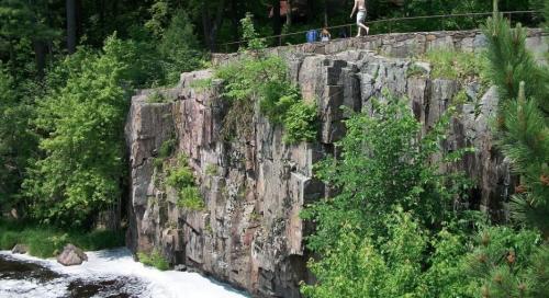 A rocky cliff covered in greenery overlooks a rushing river, with a person walking along the top path.