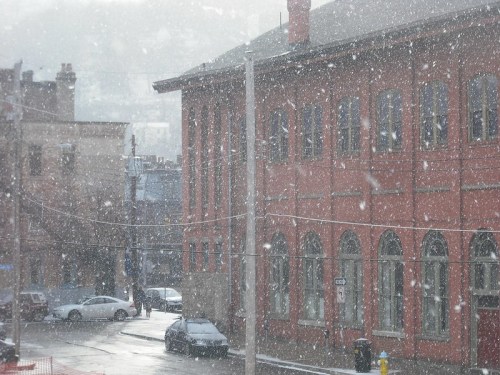 A snowy scene showing a red brick building and cars on a street, with large snowflakes falling.