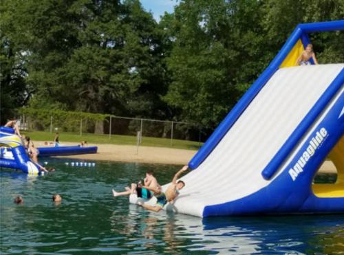 A water slide and inflatable structures at a lake, with people swimming and enjoying the sun on a warm day.