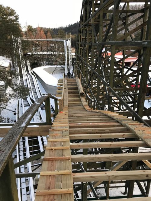 A wooden roller coaster track under construction, with a snowy landscape in the background.