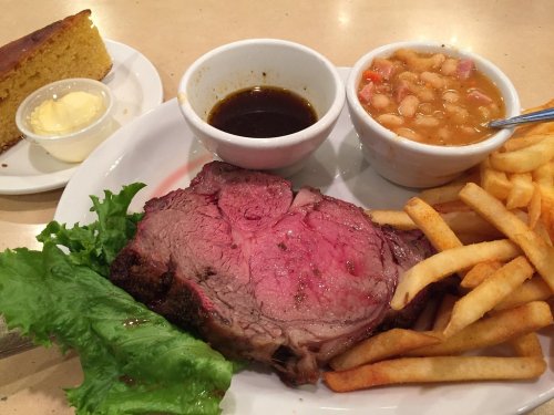 A plate featuring a juicy slice of roast beef, fries, beans, a dipping sauce, and a slice of cornbread with butter.